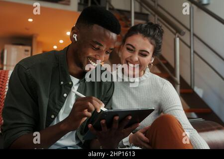 Biracial couple sitting on sofa writing on digital tablet Stock Photo