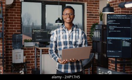 Portrait of african american programer standing working on laptop looking up and smiling at camera. Coder holding portable computer next to coworker computing big data in it agency office. Stock Photo
