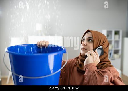 Worried Woman Calling Plumber While Collecting Water Droplets Leaking From Ceiling At Home Stock Photo