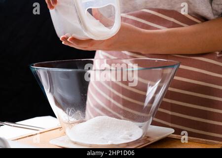Close up woman pouring flour into a mixing bowl for preparation of pastry dough on the black background. Homemade sourdough from scratch. Stock Photo
