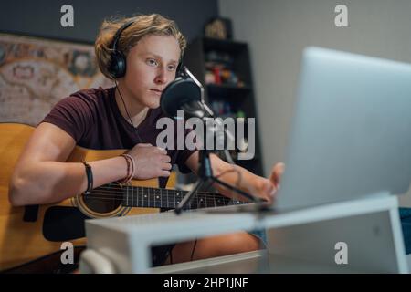 Young teenager portrait playing guitar in Headphones browsing laptop. Boy using microphone with pop filter for sound recording. Home sound studio Mode Stock Photo