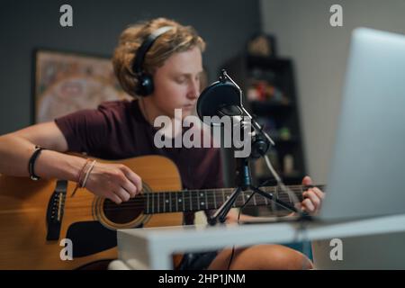 Home sound studio young teenager portrait playing guitar in Headphones with the microphone on the foreground. Modern audio recording technology concep Stock Photo