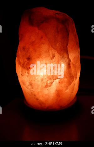 Large lit glowing dark pink Himalayan salt lamp on a wood base closeup. In a dark room is shining a glowing half a foot or so high, oblong, dark pink Stock Photo