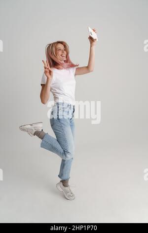 Happy young woman in casual wear jumping taking selfie holding mobile phone over gray background, full length Stock Photo