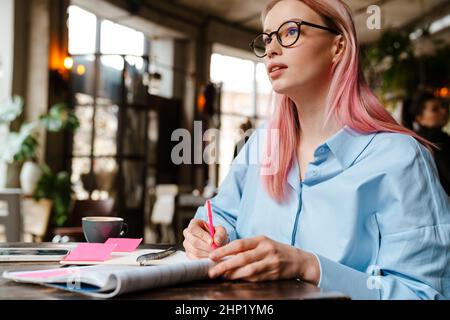 Young beautiful woman with pink hair studying and making notes while sitting in cafe Stock Photo