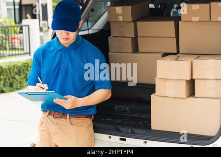 Asian young delivery man courier in uniform hold documents clipboard checking list parcel post boxes near a car for service shipment to customer, Onli Stock Photo