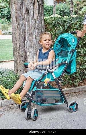 Preschooler frowns sitting in blue stroller of baby brother walking in park. Fair-haired boy sits and rests tired from long stroll on summer holiday Stock Photo