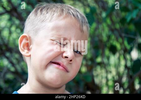 Blond preschooler grimaces making weird face expressions. Boy has fun walking outdoors in shade of trees in park on summer holiday closeup Stock Photo