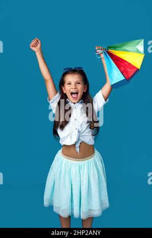 cute child girl holds many shopping bags with purchases isolated on blue background. Stock Photo