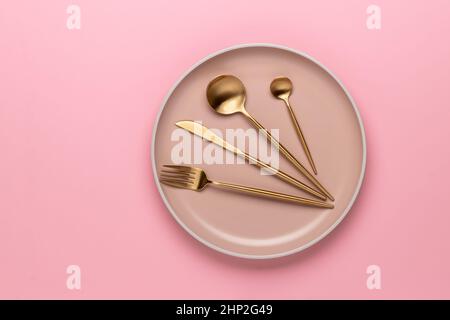 Unused beige dinner plate on a pink table, top view. Golden cutlery for serving and eating. Modern craft ceramic tableware, trendy utensils. Flat lay Stock Photo