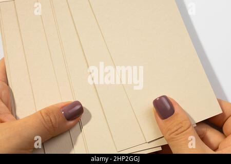 Female hands holding stack of gray-brown cardboard rectangular blank tags for clothes with shadows falling on white background. Tag mock up. Copy Stock Photo