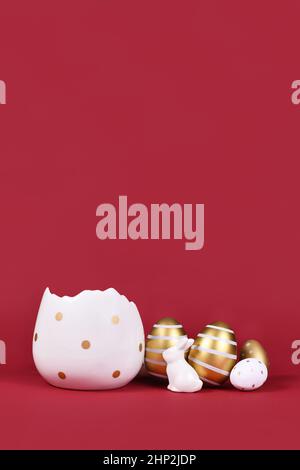 Golden and white easter eggs painted with stripes, bunny and egg cup at bottom of dark red background with copy space Stock Photo