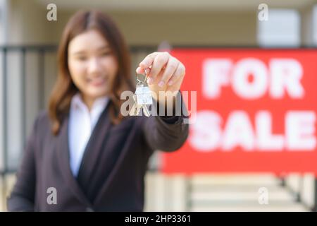 Asian real estate agent or realtor woman smiling and holding red file with showing house key in front of house. Stock Photo