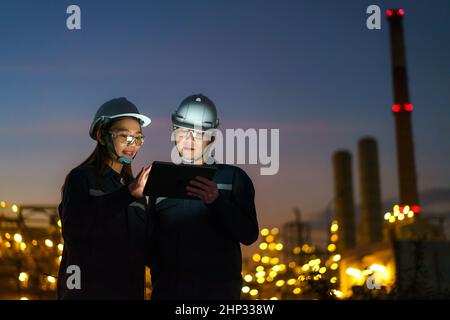 Asian engineers, man and woman are checking the maintenance of the oil refinery factory at night via digital tablets. Stock Photo