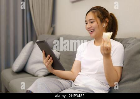 Asian woman working and meeting with her work colleague online in digital tablet and eating sandwich on sofa in living room at home at night. Stock Photo