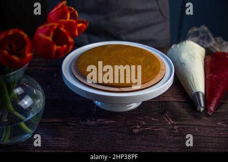 The baked cake cake lies on a white stand, pastry bags with cream and jam are next to it, a bouquet of flowers on a dark background is visible from th Stock Photo
