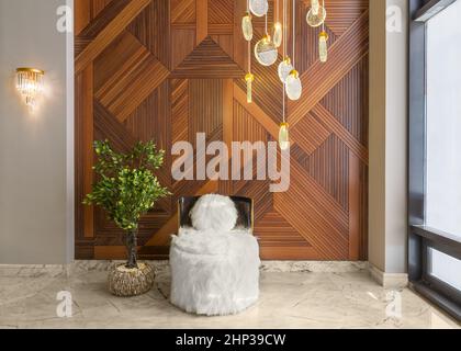 Modern white feather armless chairs, and planter with green bushes, in a hall with decorated wood cladding wall, white marble floor, and side glass wi Stock Photo