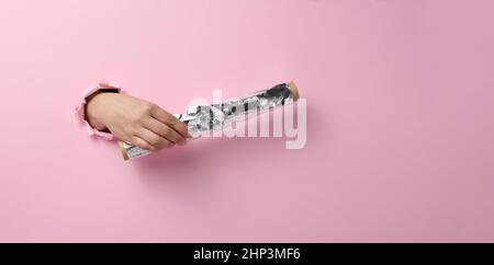 female hand holding a scroll of gray food foil on a pink background, part of the body sticking out of a torn hole. Copy space, banner Stock Photo