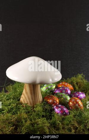 a wooden mushroom dressed in its white hat accompanying two multicolored Easter eggs on a bed of green moss Stock Photo