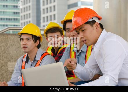 Engineering team sitting outdoors analyzing projects and checking work plan on laptop. Engineer or architec in orange helmet and holding laptop while Stock Photo