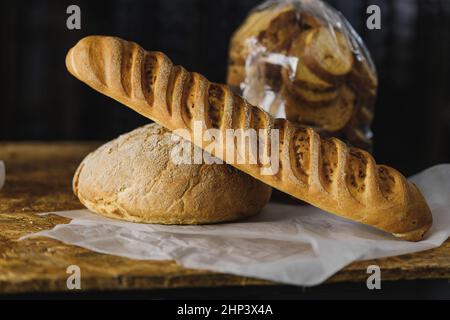 round bread and baguette bread with a bright crispy crust lies on parchment on a wooden table, dark background, side view Stock Photo