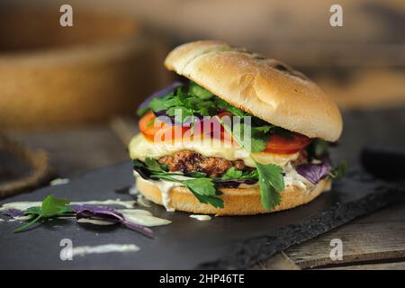 burger with beef patty, melted cheese, cream sauce, tomatoes, garnished with parsley and basil, stands on a stone blue stand, on a wooden surface, clo Stock Photo