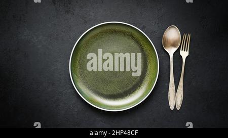 Empty ceramic plate green and old cupronickel spoon and fork on black slate table Stock Photo