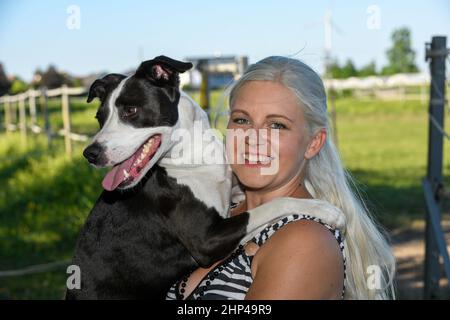 Young blonde woman with long hair holds her lovely black and white colored dog in her arms and looks to the camera. Stock Photo