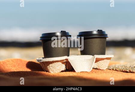 two black paper cups with coffee, covered with a lid, stand in a cardboard stand on an orange bedspread, against the background of the beach, close-up Stock Photo