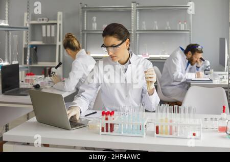 Female scientist in lab or chemist making notes on laptop by studying liquid in test tube. Stock Photo