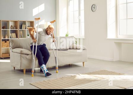 Man with leg injury feels pain and difficulty trying to get up from sofa on crutches. Stock Photo