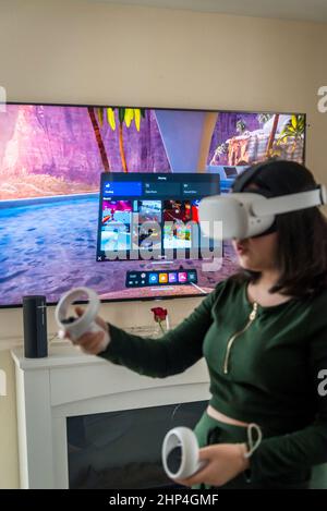 Girl with VR headset and two controllers playing with friends in virtual reality and projecting it on tv-screen behind her, London, UK Stock Photo