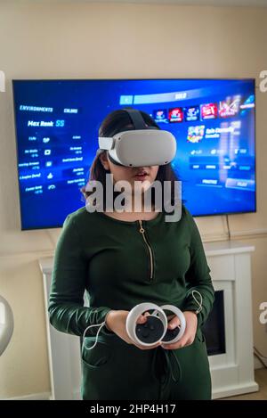 Girl with VR headset and two controllers playing with friends in virtual reality and projecting it on tv-screen behind her, London, UK Stock Photo