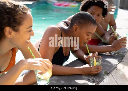 Happy biracial woman drinking from straw while enjoying sunny day with friends in swimming pool Stock Photo