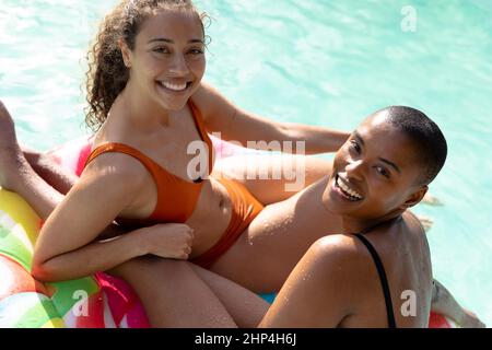 Portrait of happy young biracial women sitting together on inflatable ring in pool on sunny day Stock Photo