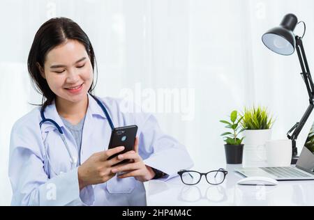 Asian doctor young beautiful woman smiling using working or holding with smart mobile phone and laptop computer at hospital desk office, technology he Stock Photo