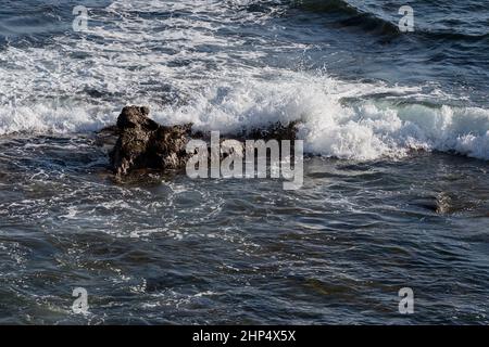 a small wave breaks over a big rock on the California coast near La Jolla with calm water in the foreground and swells and foam in the background Stock Photo
