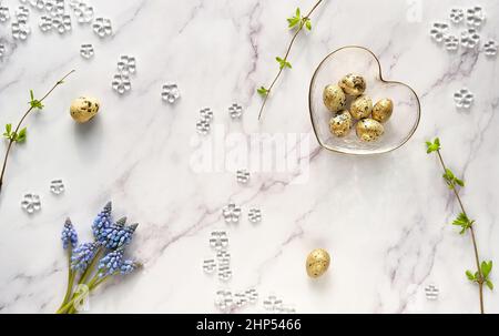 Golden quail eggs in glass heart bowl. Blue grape hyacinth flowers. Twigs with Spring leaves on off white marble. Easter off white background Stock Photo
