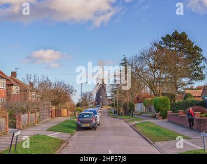 A 18th Century working windmill set in a residential street.  A road leads up to the mill and houses are on both sides. A blue sky with cloud above. Stock Photo