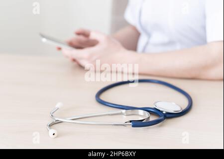Unrecognizable female doctor sits at a desk and writes a message on a smartphone. Practitioner holds a phone in his hands and answers patients Stock Photo