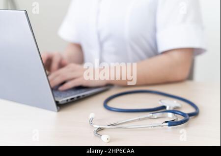 Unrecognizable female doctor in uniform uses a laptop. Faceless medical worker writes a report on the computer at the desk. Cropped. Online Stock Photo