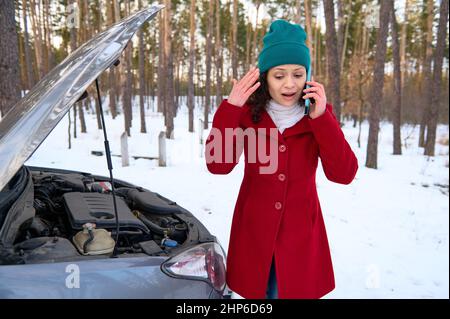 Shocked frustrated middle aged woman standing near her broken car and trying to call for roadside assistance during a car breakdown on a snowy road. A Stock Photo