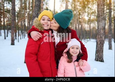 Handsome 9 years old Caucasian boy in red down jacket and yellow knit hat smiles toothy smile while his loving mom kissing him and hugging his cute yo Stock Photo