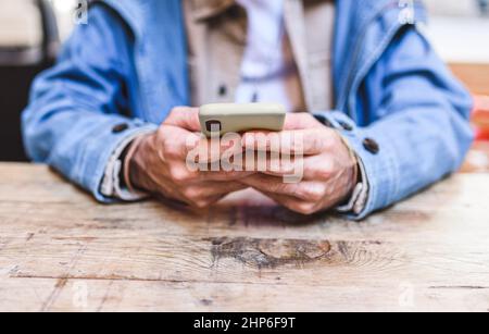 Soft focus of unrecognizable male in denim jacket browsing social media on cellphone while sitting at wooden table in cafeteria Stock Photo