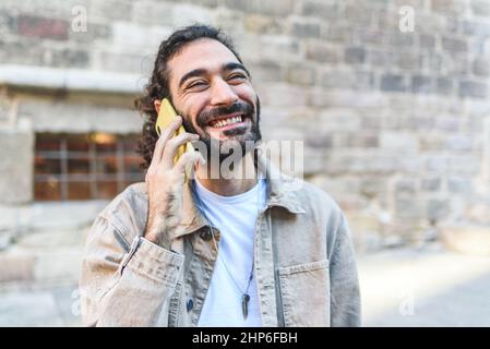 Delighted bearded male tourist with dark hair having conversation on smartphone while standing near aged stone building on street of city Stock Photo