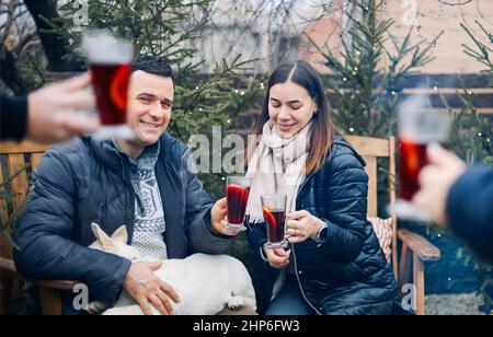 Bonfire party. Joyful young family couple gathering with friends around campfire in winter, happy people preparing and drinking hot mulled wine while Stock Photo