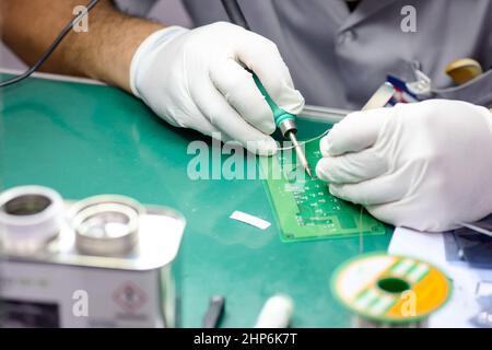Close-up technician male on rubber gloves and work suit is using soldering to weld electronics circuit board on office desk in factory Stock Photo