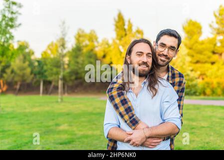 A gay couple looking at camera hugging in the park Stock Photo