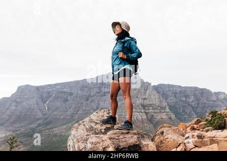 Smiling woman hiker wearing a hat enjoying the view while standing on an edge. Young female with a backpack on a hike. Stock Photo
