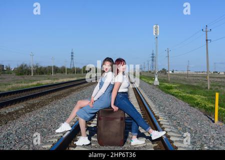 Beautiful young ladies are sitting on large suitcase on the railroad tracks. Travel concept. Stock Photo